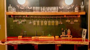 Zona de bar con taburetes, mostrador y estantes con vasos y botellas.