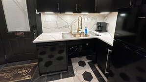 Cocina compacta con armarios oscuros, un lavamanos de acero inoxidable, una cafetera y un refrigerador negro.