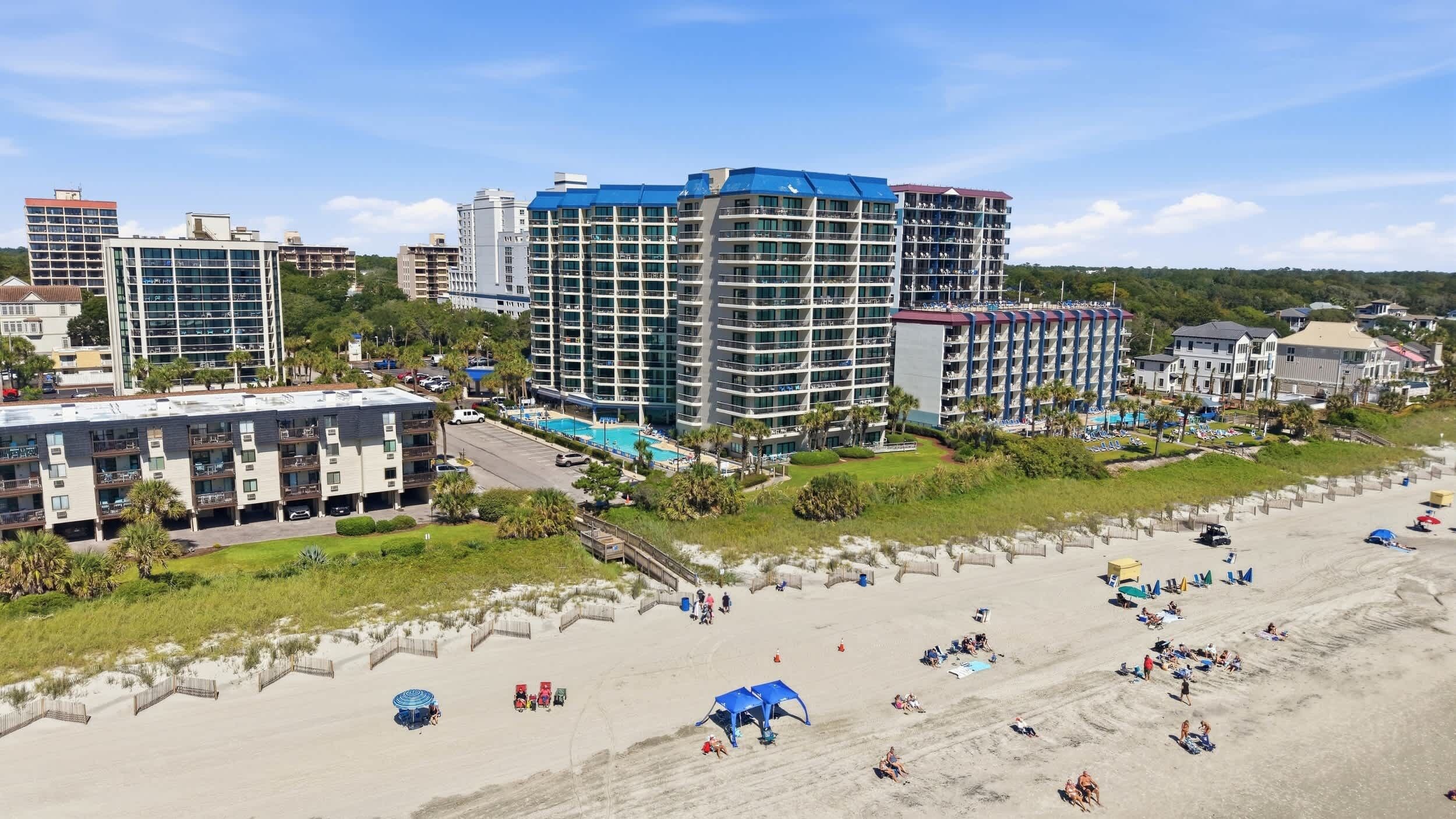 A beachfront hotel with multiple buildings, a swimming pool, and a sandy beach area with umbrellas and sun loungers.