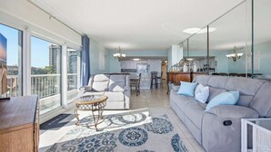 A spacious living area with a grey sofa, a small round table, and a patterned rug. A kitchen is visible in the background with modern appliances. - Serenity Suite Oceanfront w Reclining Sofa (Myrtle Beach)