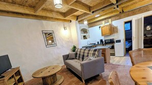 Ein gemütlicher Wohnbereich mit einer Couch, einem runden Holztisch und einem Esstisch mit Stühlen. Im Hintergrund ist eine Küche mit modernen Geräten zu sehen.
