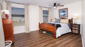 Een slaapkamer met een houten bed, een plafondventilator, een raam met jaloezieën, een nachtkastje met een lamp en een aan de muur hangend schilderij.