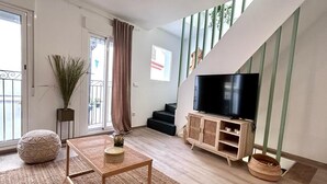 A modern living room with a TV on a wooden cabinet, a large window with curtains, a staircase, and a balcony. - Baélia Casa Sabina (Alicante)