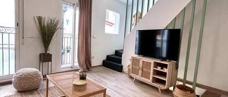 A modern living room with a TV on a wooden cabinet, a large window with curtains, a staircase, and a balcony.