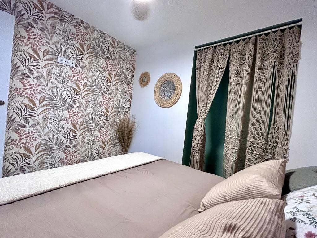 Une chambre à coucher avec un lit, un papier peint à motifs, des rideaux en macramé et une assiette décorative accrochée au mur.