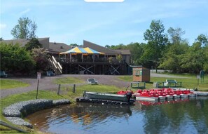 A lakeside area with a dock, a boat, and a building with a colorful awning. - Poconos Basecamp | Pickleball, Hot Tub & Sauna (Albrightsville)
