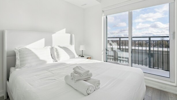 A modern hotel room with a large bed, a nightstand, a window with a view of the city, and a balcony with a railing. - Home Richmond Hill 2bed2.5bath | Parking (Richmond Hill)