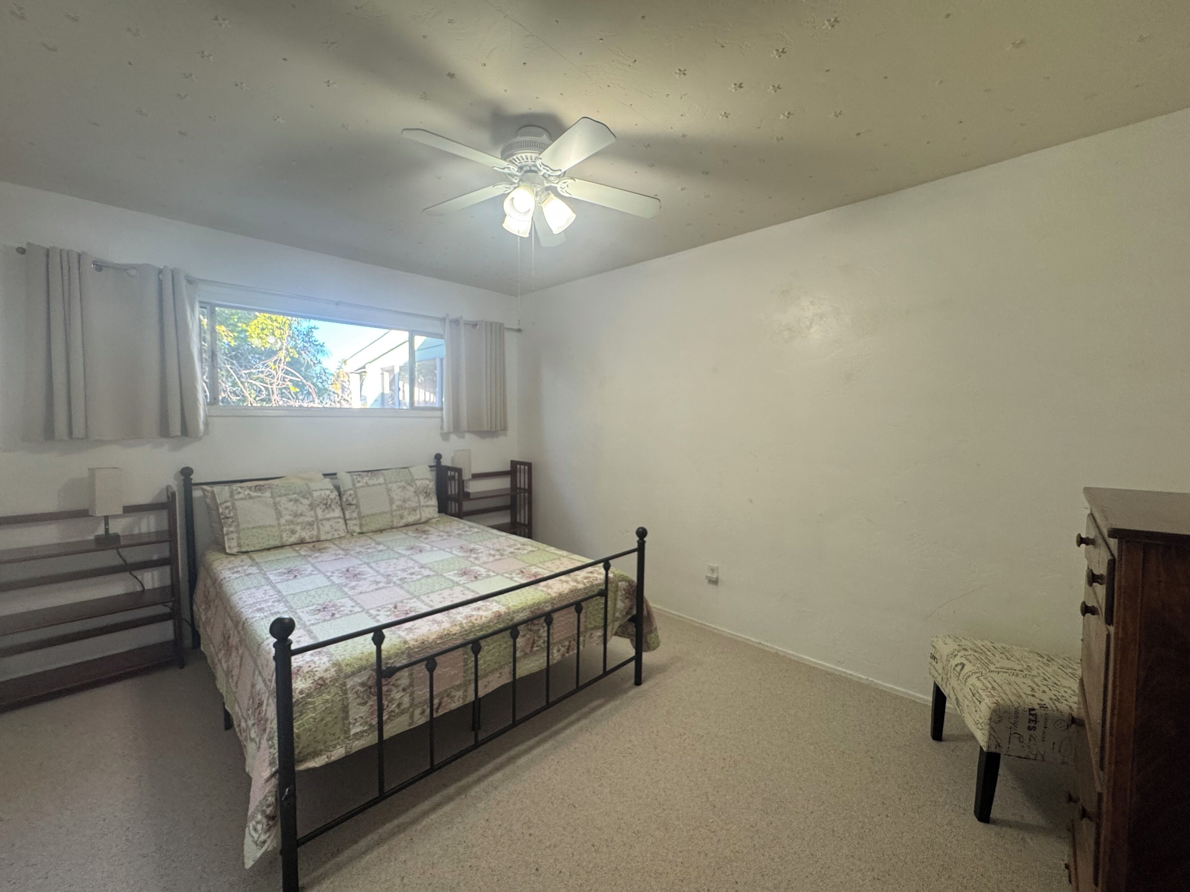A bedroom with a single bed, a ceiling fan, a window with curtains, a small bench, and a wooden cabinet.