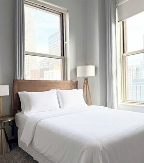 A hotel room with a large bed, a nightstand, a lamp, and a view of buildings through the window.