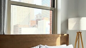 A hotel room with a bed, a window with a city view, and a floor lamp.