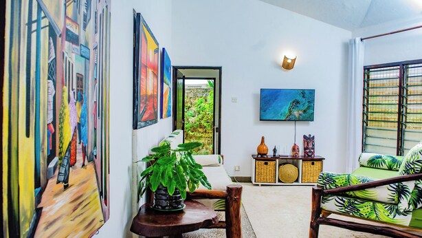 Ein Zimmer mit Holzstuhl, einem Tisch mit Blumentopf, einer Couch mit grüner Kissen und einem an der Wand montierten Fernseher. Eine Tür führt in einen Garten.