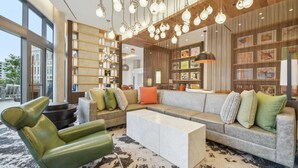 Ein modernes Wohnzimmer mit einem Ecksofa, einem grünen Sessel und einem Couchtisch. Hängende Leuchten und ein Bücherregal mit diversen Gegenständen sind ebenfalls vorhanden.