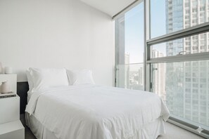 Une chambre d’hôtel moderne avec un grand lit, une table de chevet avec une lampe et une vue sur la ville grâce à de grandes fenêtres du sol au plafond.