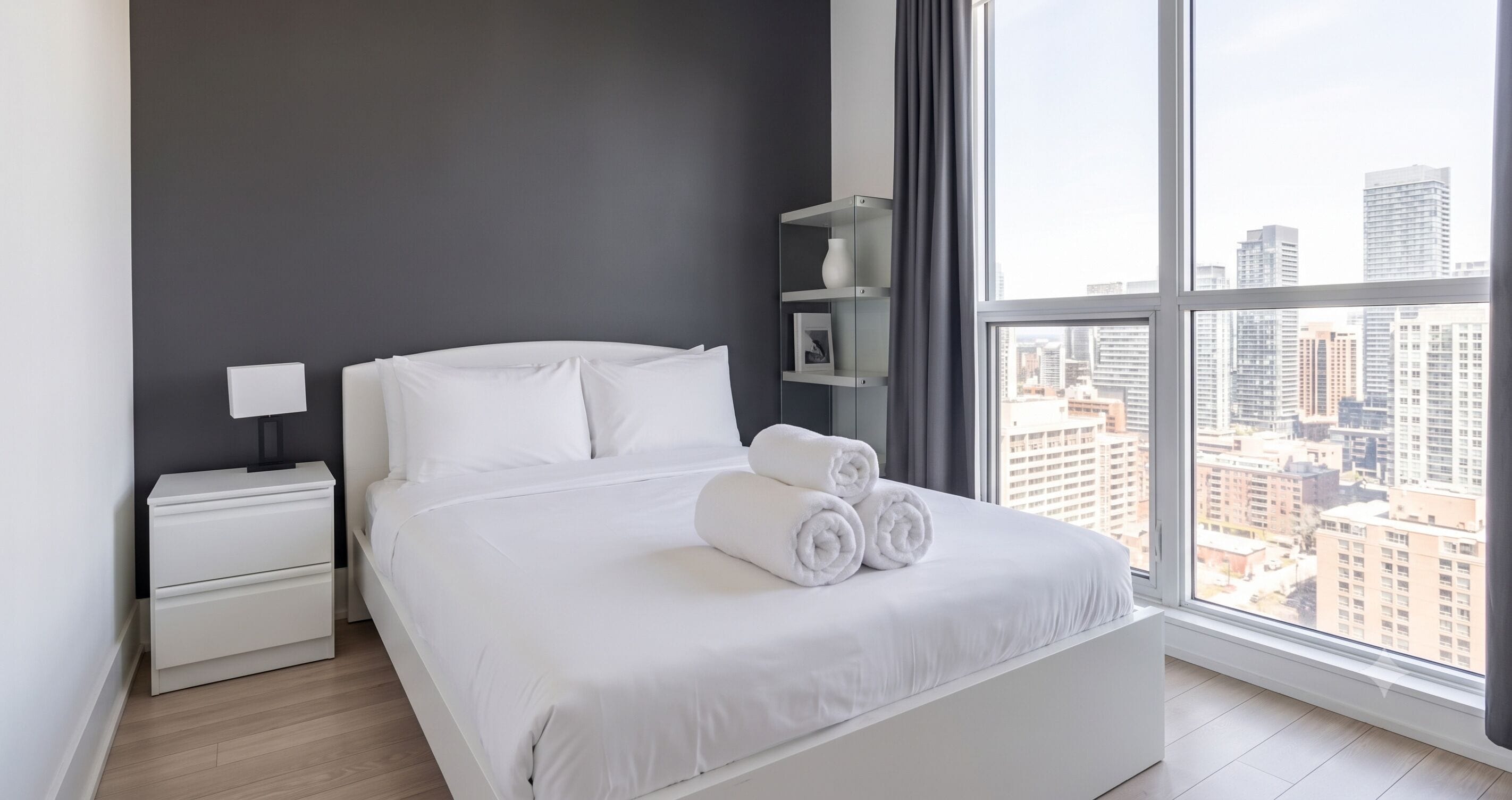 A modern hotel room with a large window, a neatly made bed, a bedside table, and a small shelf with a picture frame.