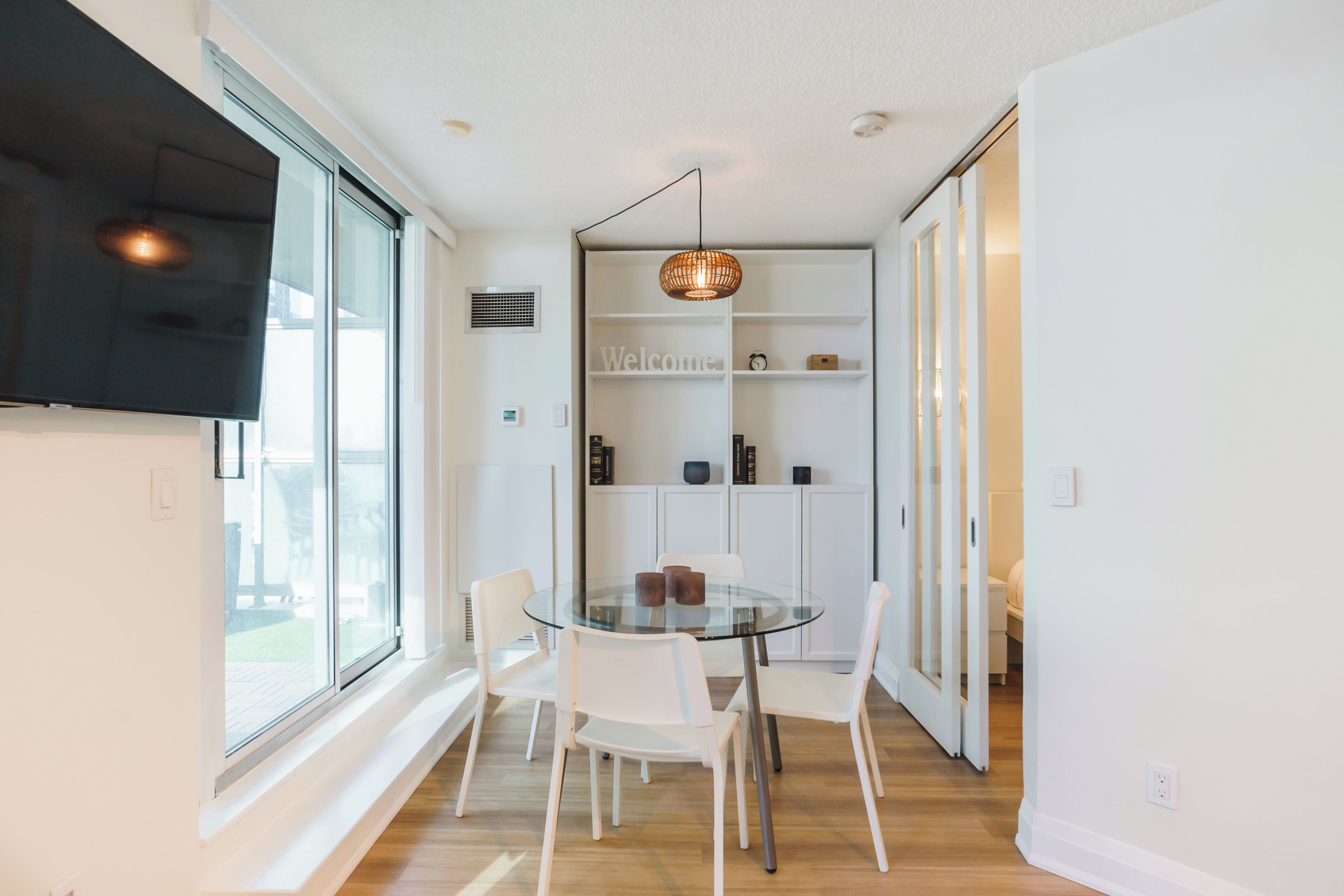 A modern dining area with a glass table, white chairs, a hanging light fixture, and a white cabinet with shelves.