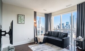 Un salon moderne doté d’une grande fenêtre, d’un canapé gris, d’un téléviseur à écran plat fixé au mur et offrant une vue sur la ville.