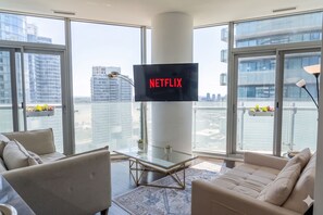 A modern living room with a large window, a flat-screen TV mounted on the wall, and a glass coffee table. - StaysToronto (Toronto)