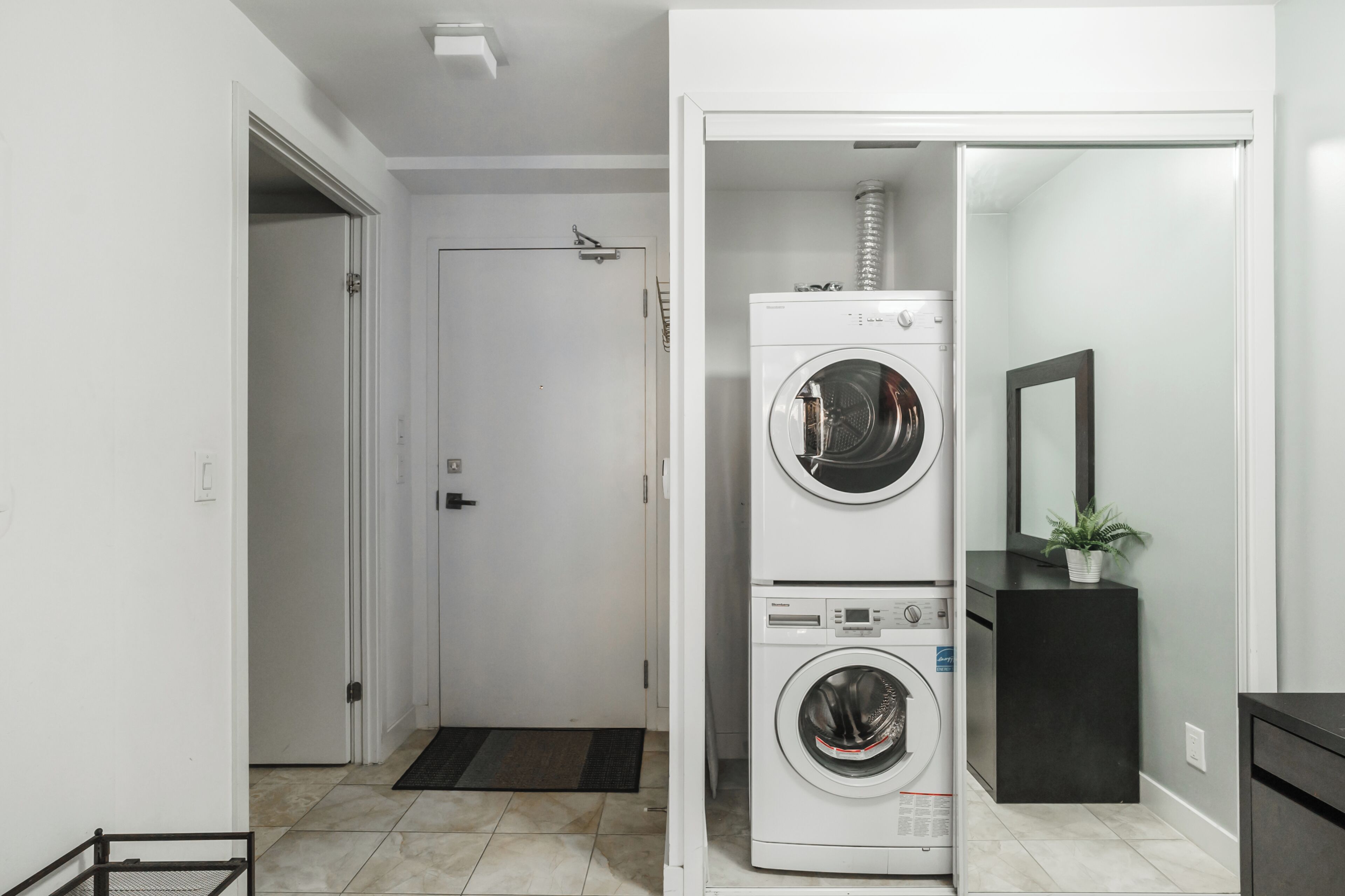 A laundry room with two stacked washing machines, a door, a small plant on a black cabinet, and a tiled floor.