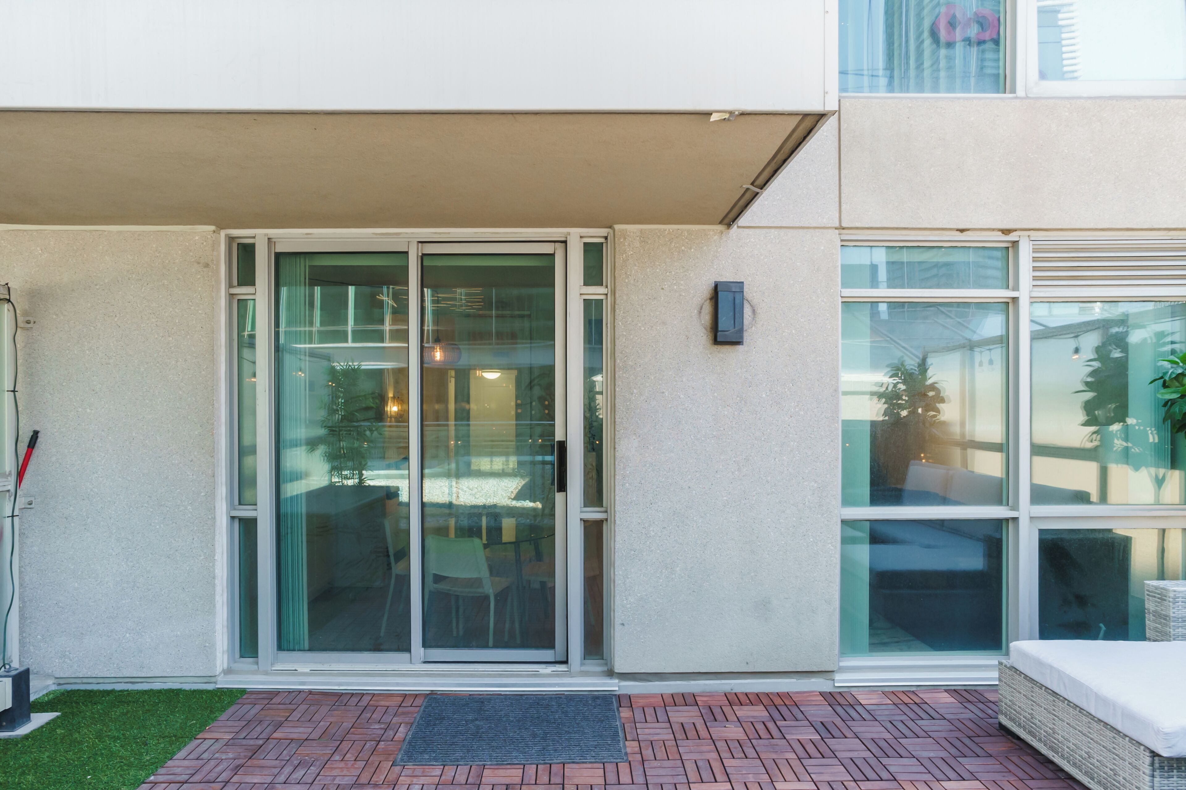 Une entrée d’immeuble avec une porte en verre, un petit espace extérieur aménagé avec des sièges et un pot de fleurs.