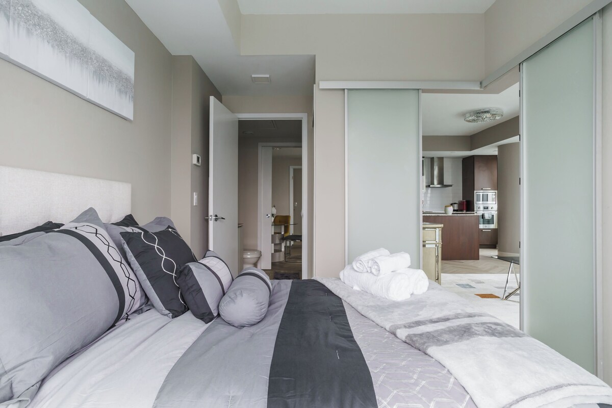 A modern hotel room with a neatly made bed, a bathroom with a toilet and sink, and a kitchenette in the background.