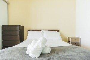 Een net opgemaakt bed met witte lakens en een grijze deken, een houten dressoir links en een klein nachtkastje met lades rechts.