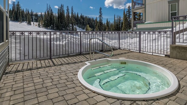 Een hottub op een geplaveid terras met uitzicht op een besneewd berglandschap en een gebouw op de achtergrond.