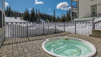 A hot tub on a paved patio with a view of a snowy mountain landscape and a building in the background.