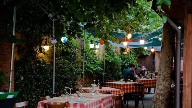 An outdoor dining area with tables covered in checkered tablecloths, surrounded by greenery and string lights. - The Remembrances Suite (Milan)