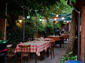 An outdoor dining area with tables covered in checkered tablecloths, surrounded by greenery and string lights. - The Remembrances Suite (Milan)