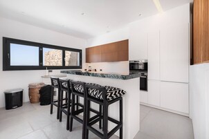 Una cocina moderna con armarios blancos, encimera de mármol y horno empotrado. Hay tres taburetes negros con cojines estampados.