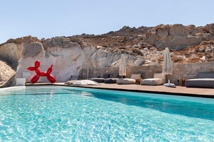 Una piscina con una escultura roja, sillas de descanso y un paisaje montañoso rocoso al fondo.