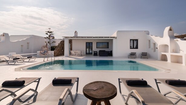 Un área de piscina en la azotea con sillones reclinables, una mesa de madera y un edificio de paredes blancas y chimenea.