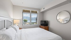 A bedroom with a bed, a TV on a wooden cabinet, a window with blinds, and a round mirror on the wall. - Lakefront Resort Condo with Pool (West Kelowna)