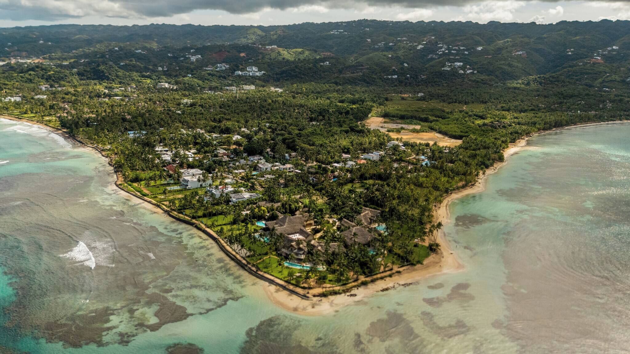 An aerial view of a coastal resort with multiple buildings, a beach, and surrounding greenery.