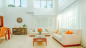 A spacious living room with a sofa, coffee table, and a wooden chair. There is a decorative clock on the wall and a window with curtains. - Lux Villa Playa Bonita (Las Terrenas)