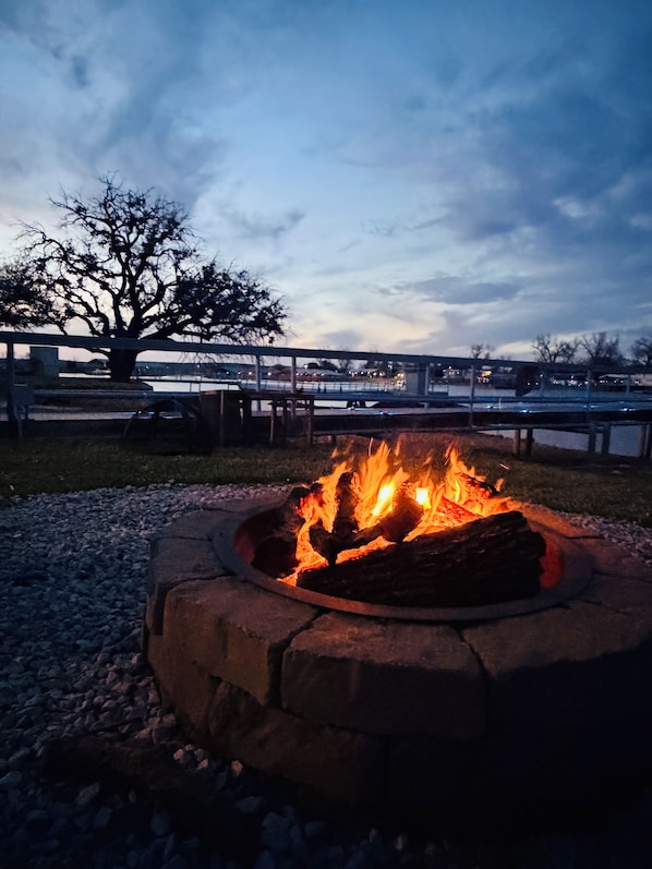 A fire pit with logs, surrounded by gravel, with a tree and a fence in the background.