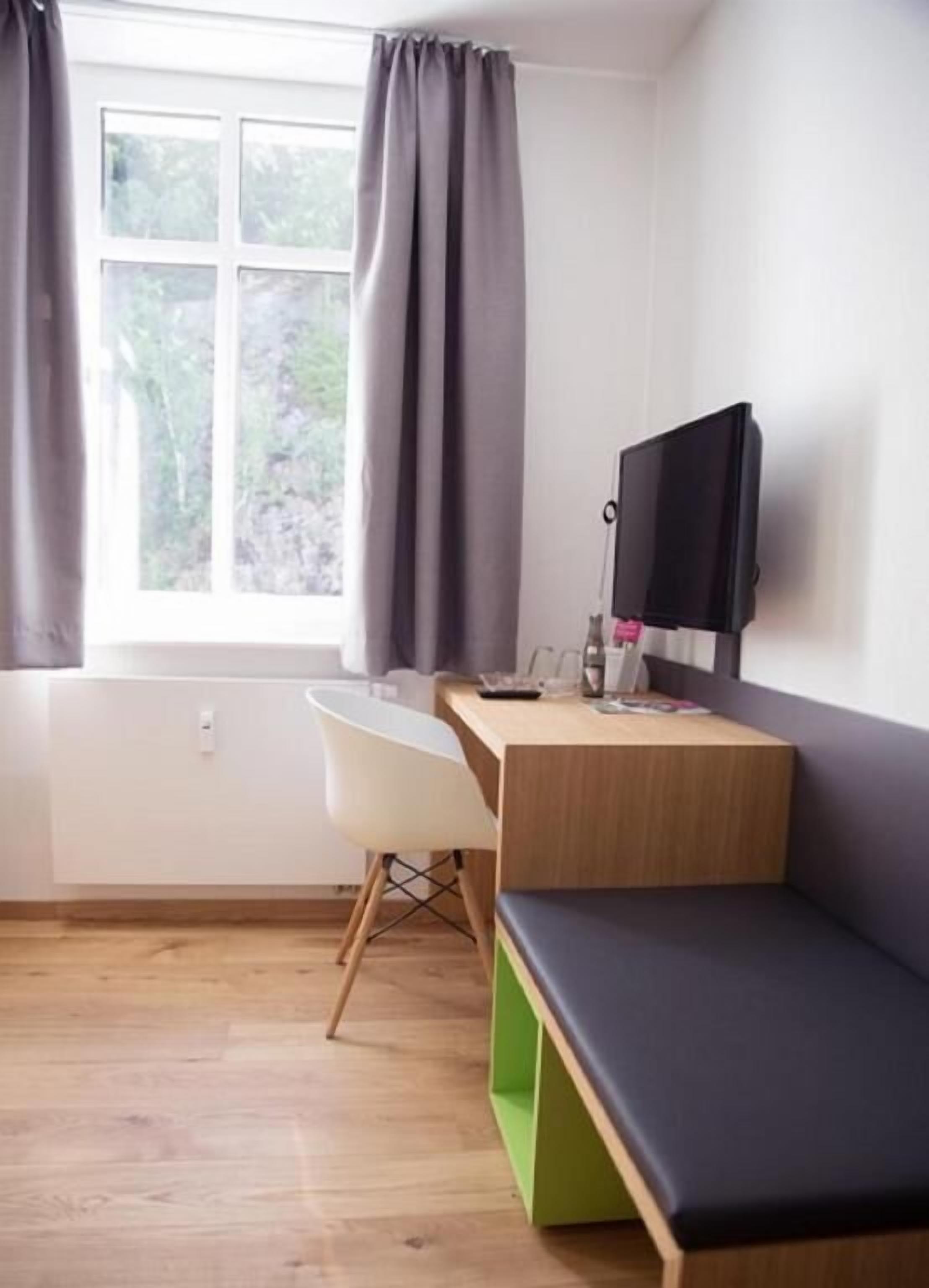 Quarto com escrivaninha de madeira, uma cadeira, um banco, uma televisão e janela com cortinas.