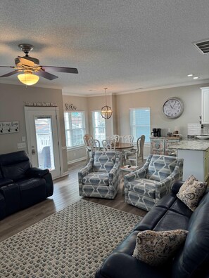 A living room with a black leather sofa, patterned armchairs, a dining table, and a kitchen area with a clock on the wall. - Ocean Lakes 1383 | Spacious 5-Bdrm,3 bath Sleeps 13 (Myrtle Beach)
