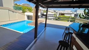 A rooftop pool with a wooden deck, a covered seating area with stools, and a view of surrounding buildings. - Luxury House in Gated Community in Metropolitan Area (Mogi das Cruzes)
