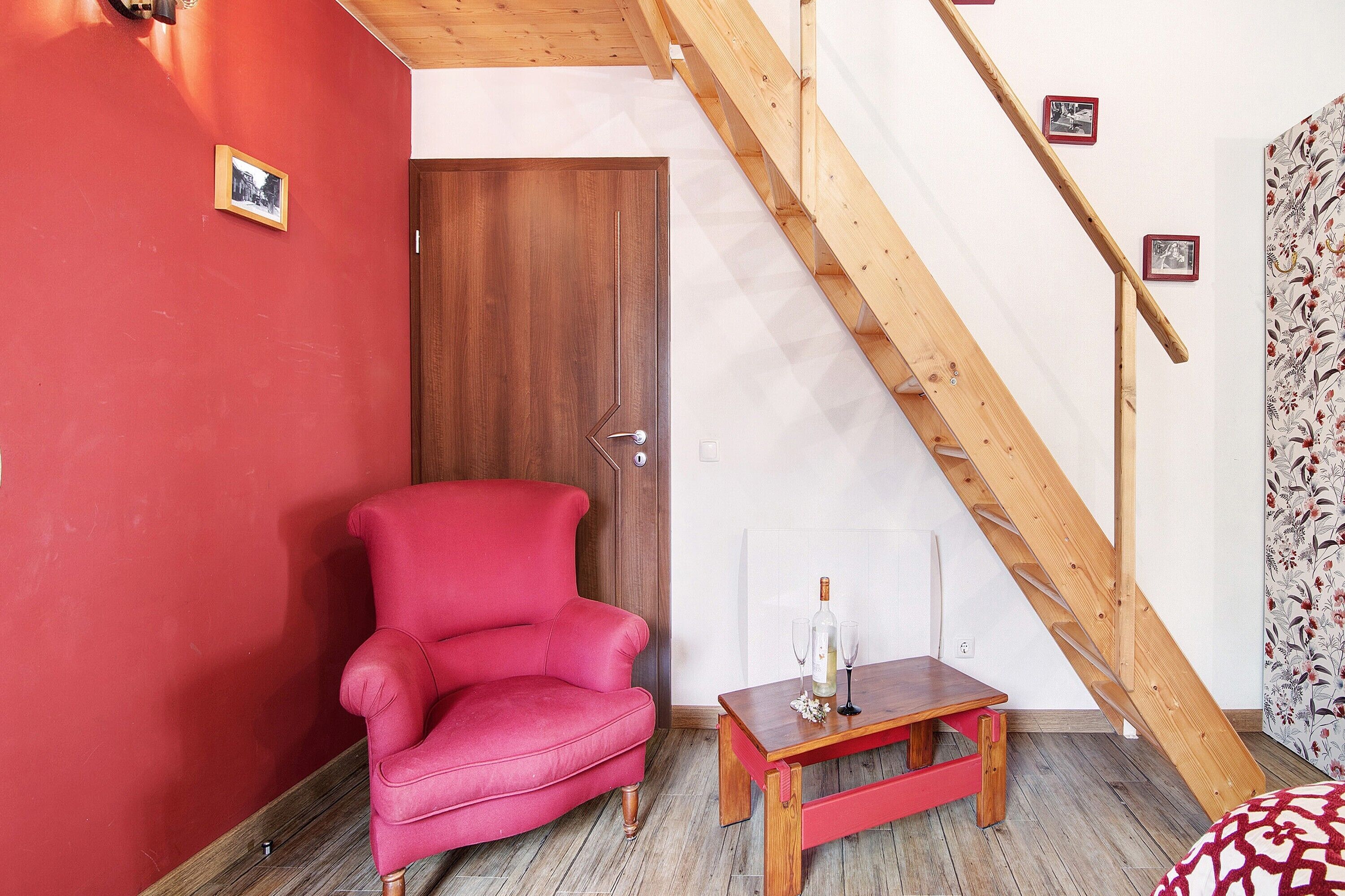 A room with a red wall, a wooden door, a red armchair, a wooden table with a bottle and glasses, and a wooden staircase.