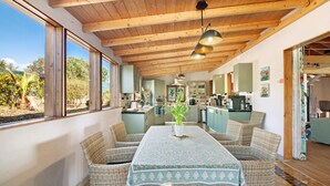 A dining area with wicker furniture, a table with a patterned tablecloth, and a view of the kitchen and garden. - Venice Room – Naturist B&B for Couples (Monte da Charneca)