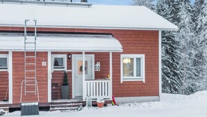 Punainen puutalo, jossa on valkoinen kuisti, ovi ja ikkuna. Talon seinää vasten on nojaamassa tikapuut, ja kuistilla on ruukussa oleva kasvi.