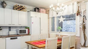 Kompakti keittiö, jossa on valkoiset kaapit, mikroaaltouuni ja jääkaappi. Keittiössä on ruokapöytä tuoleineen ja pieni valkoinen, pehmeä jakkara. Ikkunasta avautuu näkymä lumiseen maisemaan.