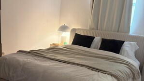 A neatly made bed with a white duvet and a beige throw blanket, a bedside lamp, and a window with curtains. - Beautiful, cozy boho chic apartment in the Heart of South Beach, Miami (Miami Beach)