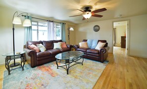 A living room with a brown leather sofa, two glass coffee tables, a ceiling fan, and a large window with curtains. - 1014b Swope (Colorado Springs)