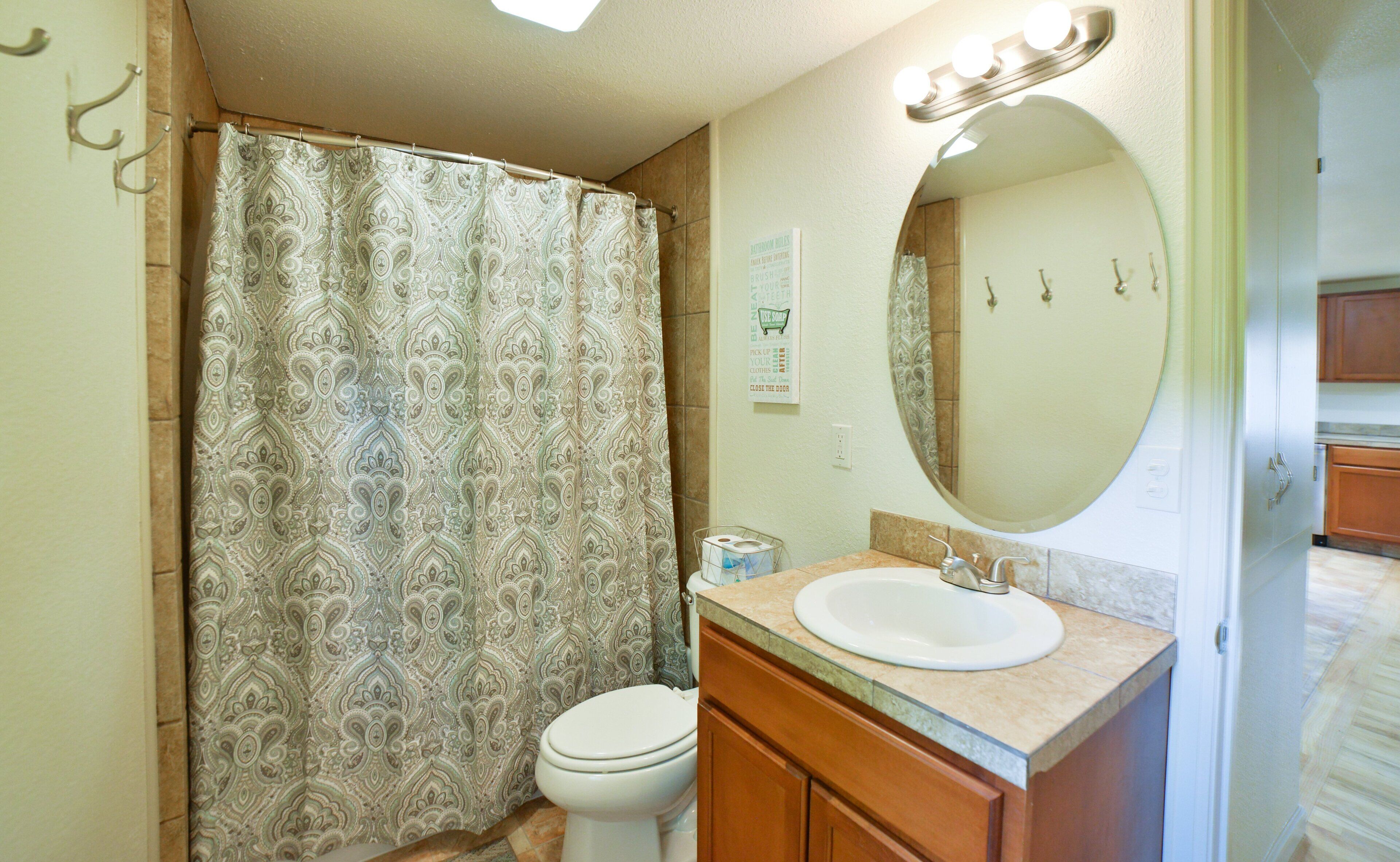 A bathroom with a patterned shower curtain, a toilet, a sink with a mirror, and a sign on the wall.