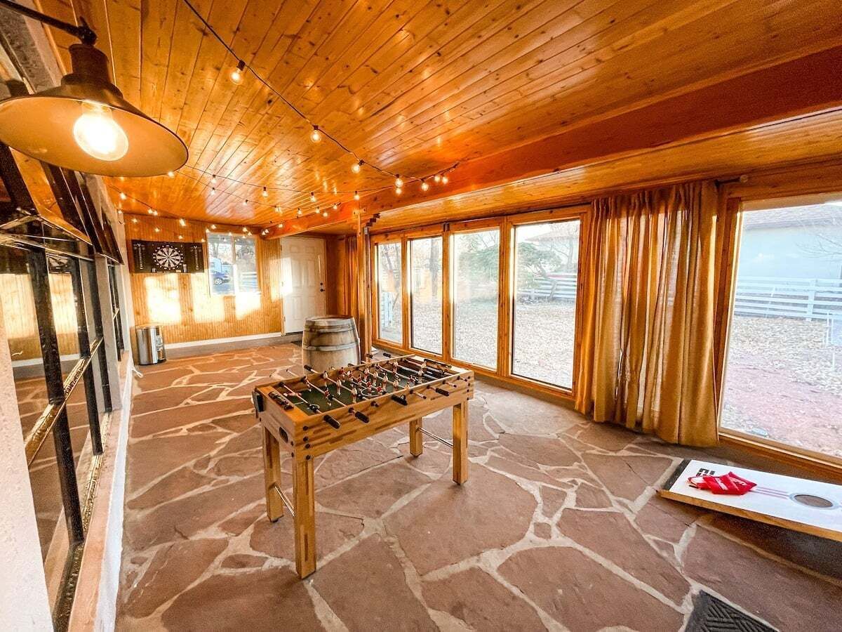 A room with a foosball table, stone flooring, wooden ceiling, large windows, and a view outside.