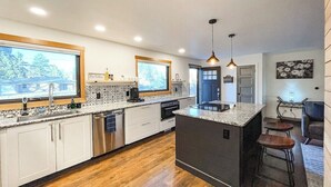 A modern kitchen with white cabinets, a large island with stools, a built-in oven, and a window overlooking the backyard. - 1625 E Cache La Poudre (Colorado Springs)