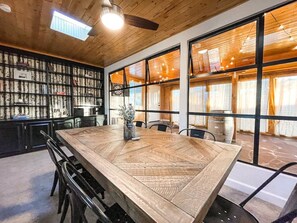 Una amplia mesa de comedor de madera con sillas, una estantería y ventanas grandes con cortinas.