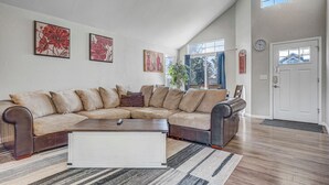 Ein geräumiges Wohnzimmer mit einer großen, L-förmigen Eckcouch, einem weißen Couchtisch und einem Streifenmuster-Teppich. Zwei große Fenster sorgen für natürliches Licht und bieten einen Blick ins Freie.